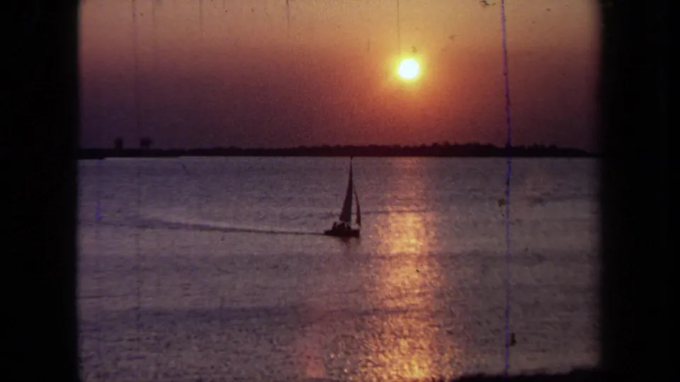 1971 California 8mm sunset frame with a distant sailboat silhouetted below a warm sun over the water.