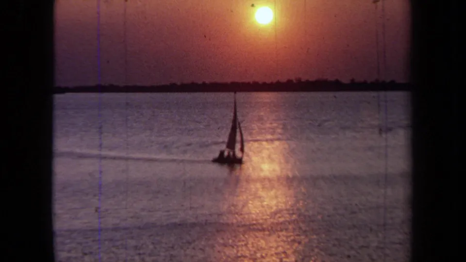 1971 California home-movie frame showing a sailboat silhouetted against reflective sunset water.