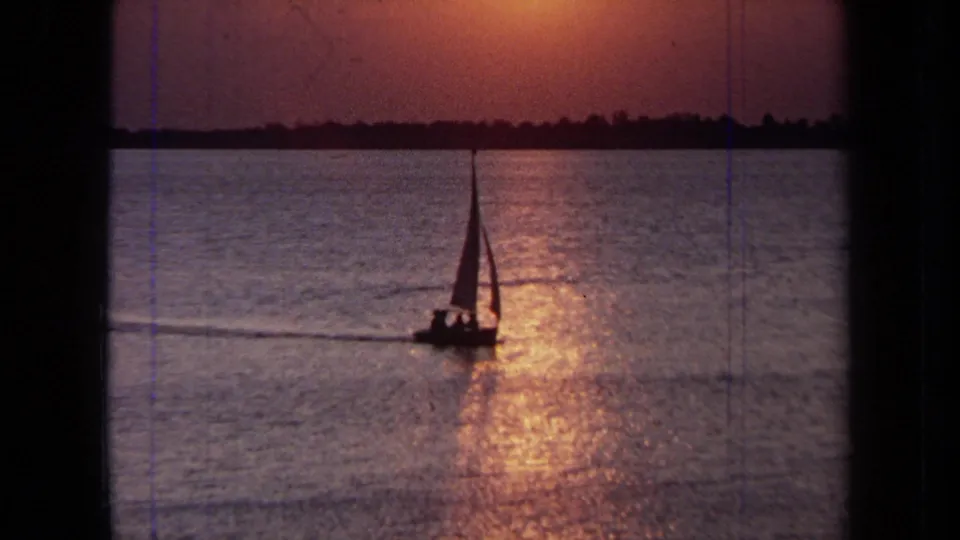 1971 California 8mm frame of a small sailboat silhouetted on calm water beneath a warm sunset reflection.