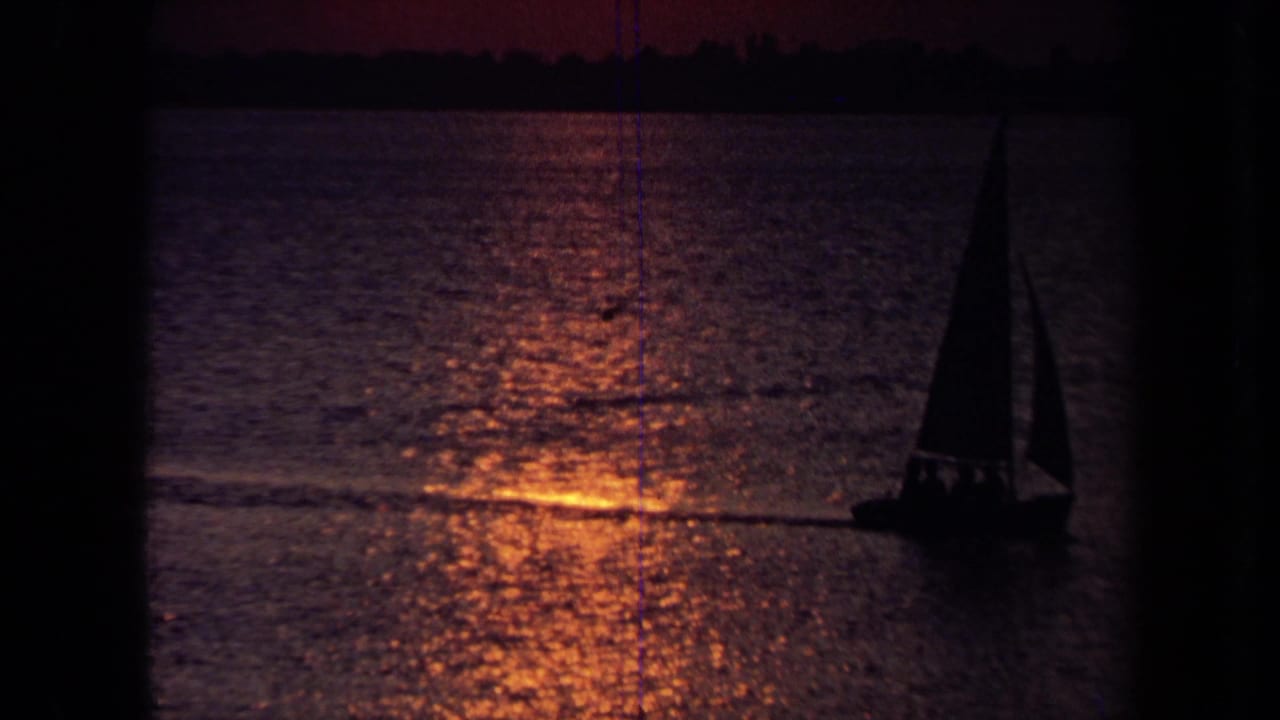 Sailboat with dark sail on orange-purple water near a distant shoreline.