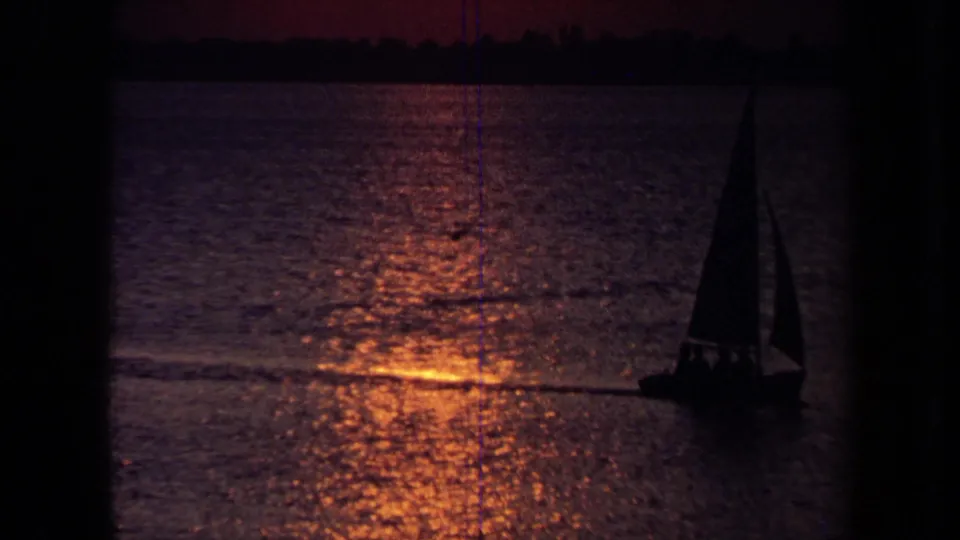 1971 California 8mm frame with a sailboat silhouetted against dusk water and a low dark shoreline.