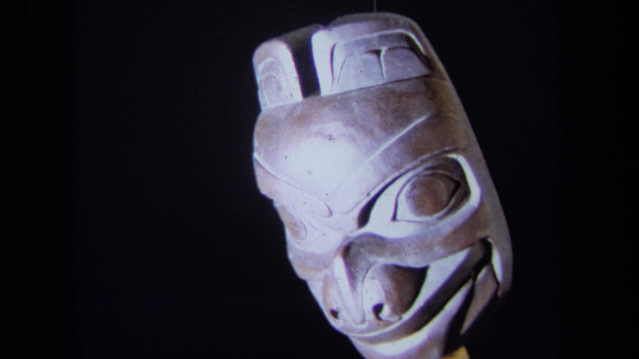 1971 8mm frame of a carved mask in close-up against a dark display background, with stylized eye, brow, and mouth forms visible.