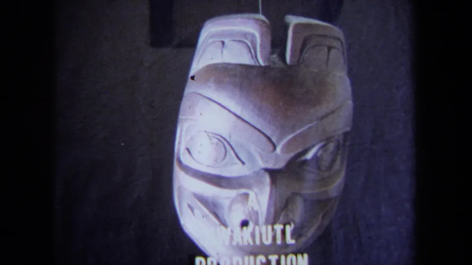 Front-facing close-up of the carved mask with white "KWAKIUTL PRODUCTION" text visible along the lower edge of the frame.