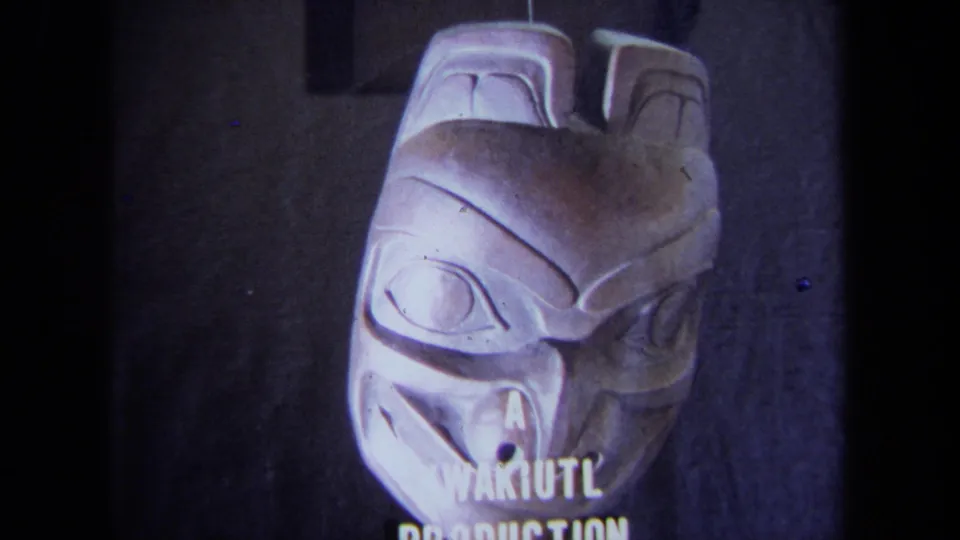 1971 California 8mm frame showing a pale carved mask with stylized eyes and partial text reading “A KWAKIUTL PRODUCTION.”
