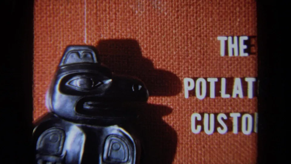 1971 8mm frame showing a wider view of the carved bird figure with cropped white text reading parts of The Potlatch Customs.