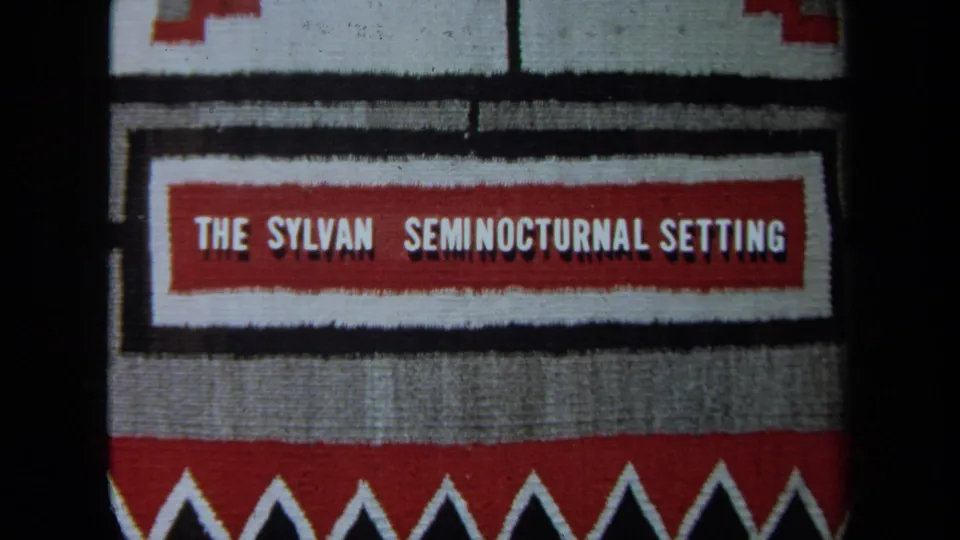 1971 California 8mm title card reading “THE SYLVAN SEMINOCTURNAL SETTING,” set on red, white, black, and gray patterned fabric.