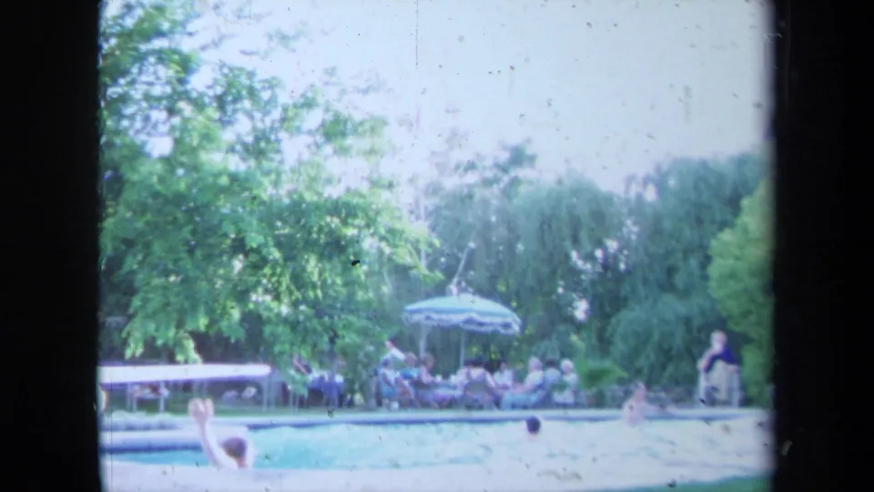 1971 California 8mm poolside scene with distant people swimming and lounging near chairs, an umbrella, and leafy trees.