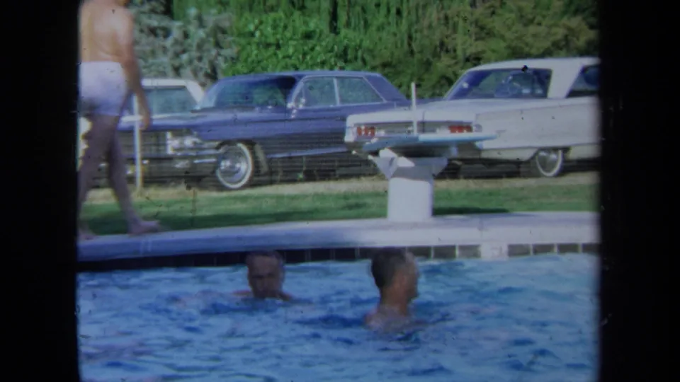 Two adult men swim in a pool with large period sedans parked on the grass behind them in California, 1971.