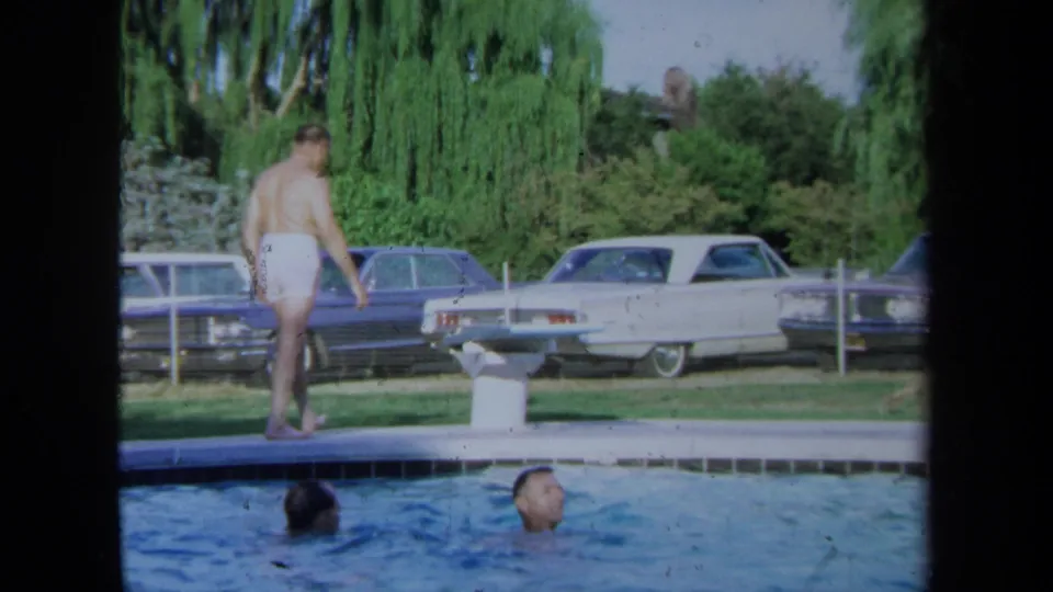An adult man in white swim trunks walks beside the pool while two men swim, with parked cars in the background.