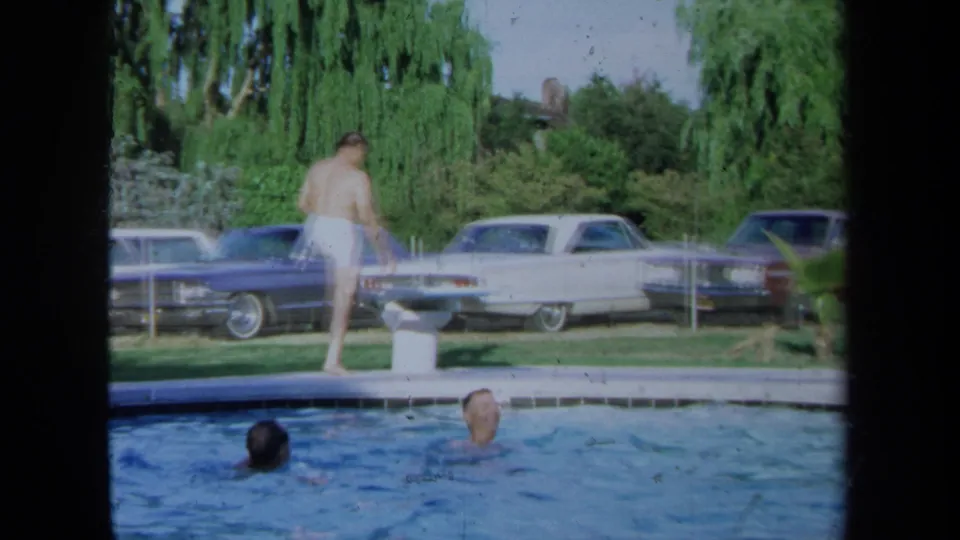 Poolside view of an adult man walking in white swim trunks as two swimmers face the edge, with 1970s cars behind them.