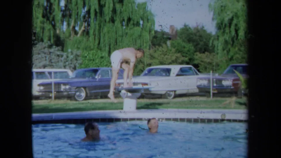 In a 1971 California backyard pool scene, a man stands on a diving board while adults swim below and cars are parked beyond the fence.