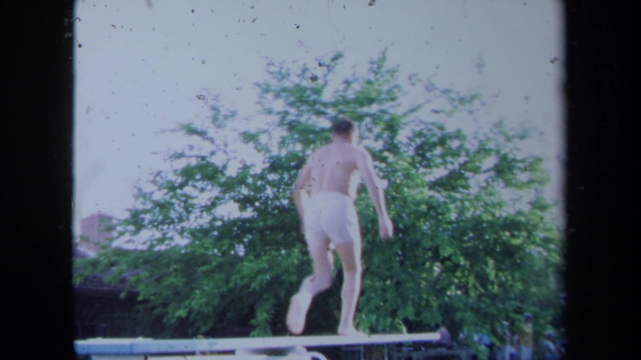Man runs along a diving board with leafy trees behind him.