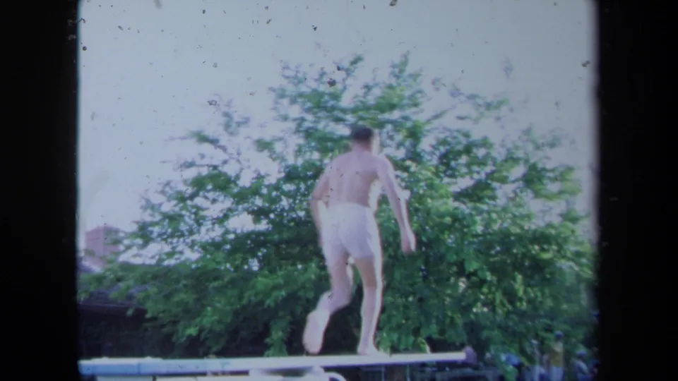 The man moves along the backyard diving board in white swim trunks, framed by green trees and bright 8mm exposure.