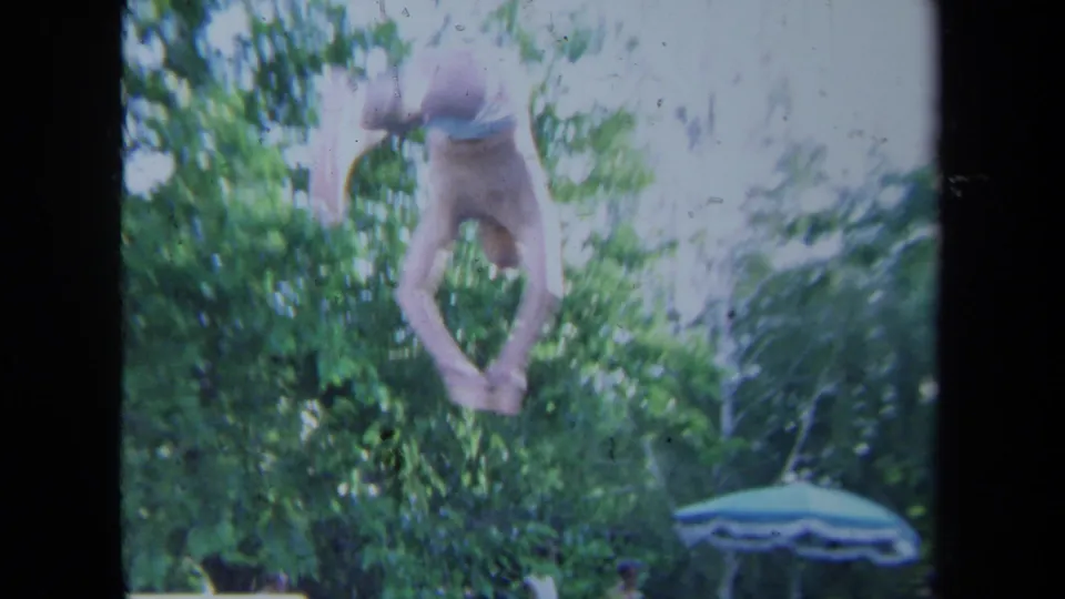 The diver is captured midair during a backyard pool jump, with leafy landscaping and a striped patio umbrella below.