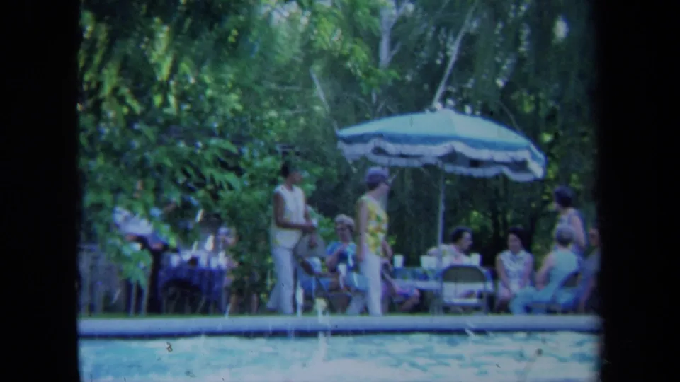 Adults sit and stand around a backyard pool in California, with a striped patio umbrella and dense greenery behind them.