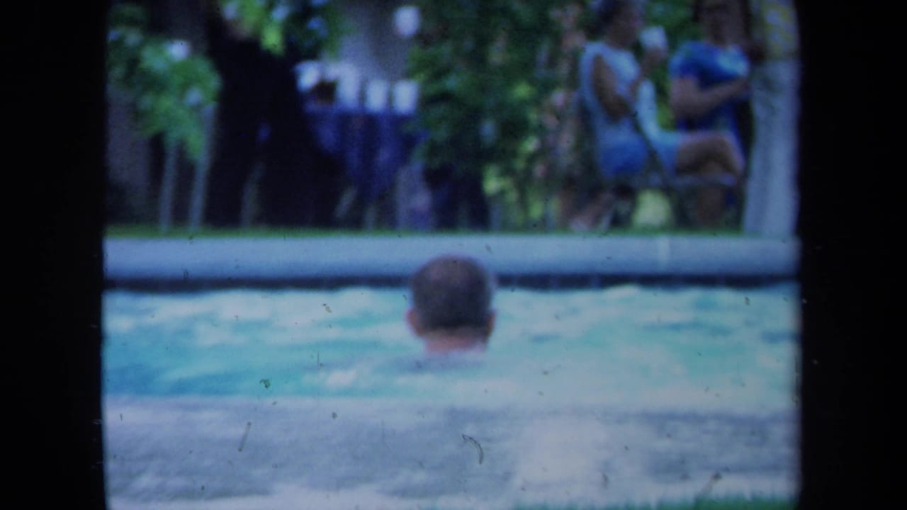 1971 California home-movie frame with a swimmer's head above the pool and seated adults beyond the water.
