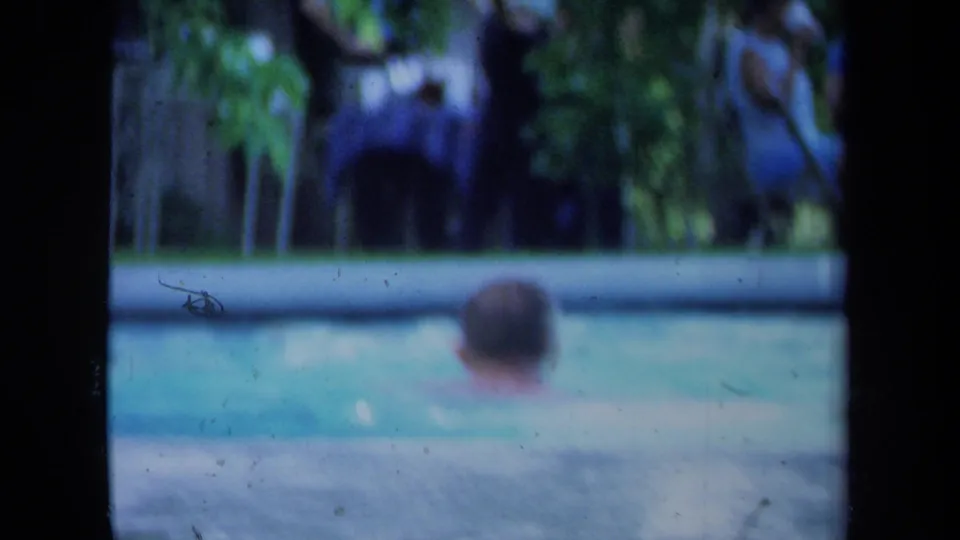 Soft 8mm frame from a 1971 California backyard pool scene, with a swimmer in the foreground and adults behind.