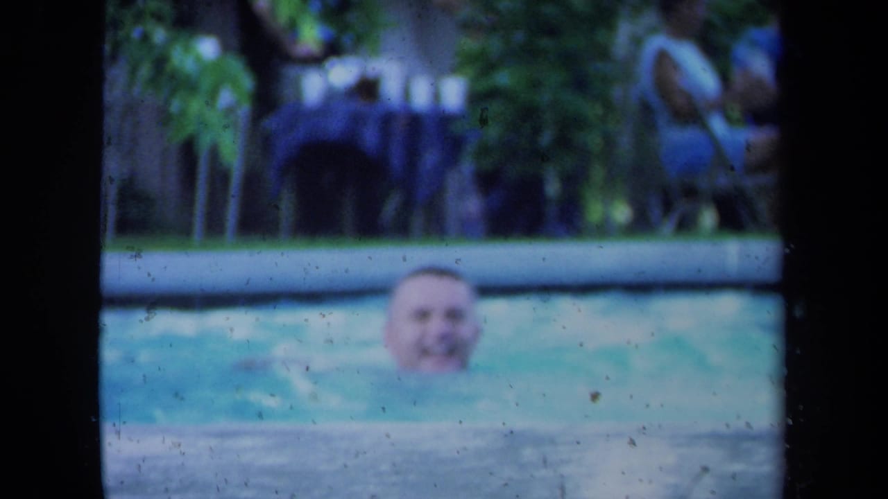 Adult man smiles from the water in a California pool scene, with trees and soft 8mm color in the background.