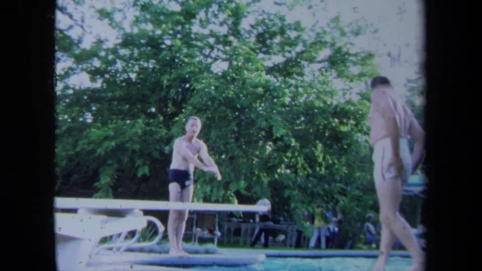 Two adult men stand near a pool and diving board in California, one on or near the board and the other at poolside.