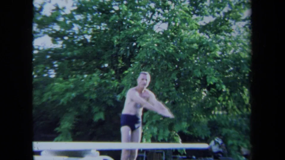 Adult man in swim trunks gestures beside a diving board in the 1971 California poolside scene.