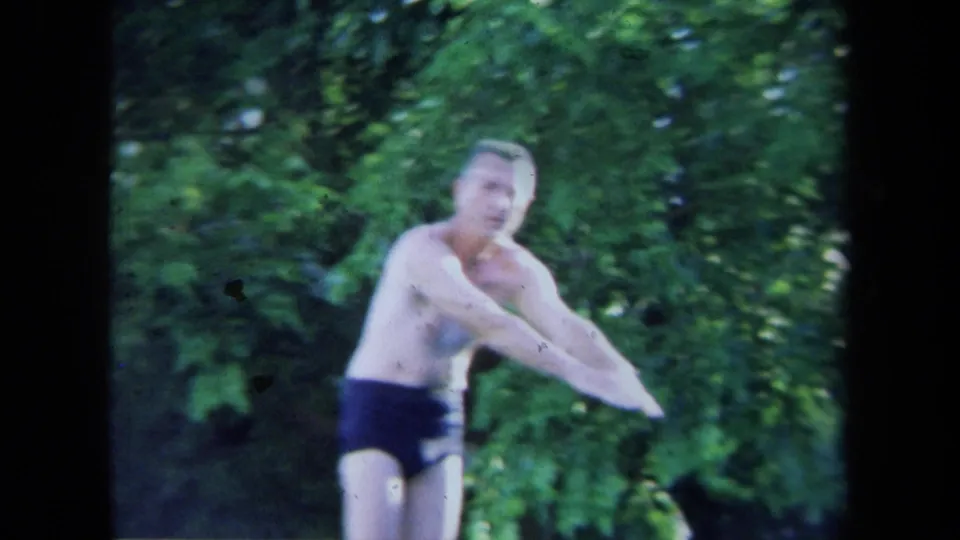 Adult man in dark swim briefs prepares to dive in 1971 California home-movie footage, with green trees behind him.