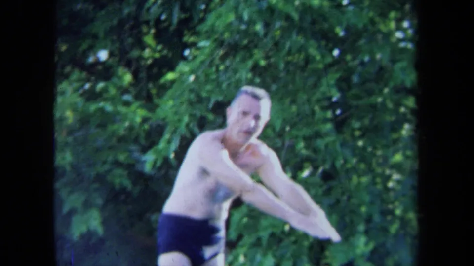 The diver leans forward in dark swim briefs, his face visible against leafy California poolside greenery.