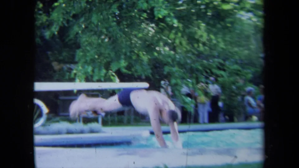 The man is captured mid-dive from a white board into pool water, with motion blur and trees in the 1971 California scene.