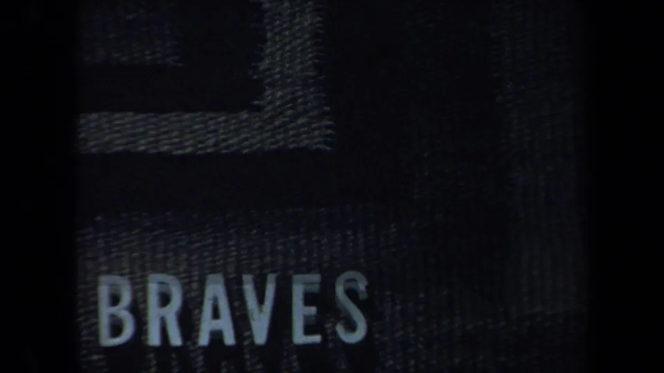 A dark, grainy frame shows a textured bag or case with the word BRAVES, creating a visible brand or team-name clearance note.