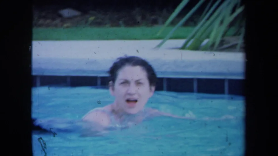 In this 1971 California Super 8 frame, a woman faces the camera from blue pool water near a tiled pool edge.