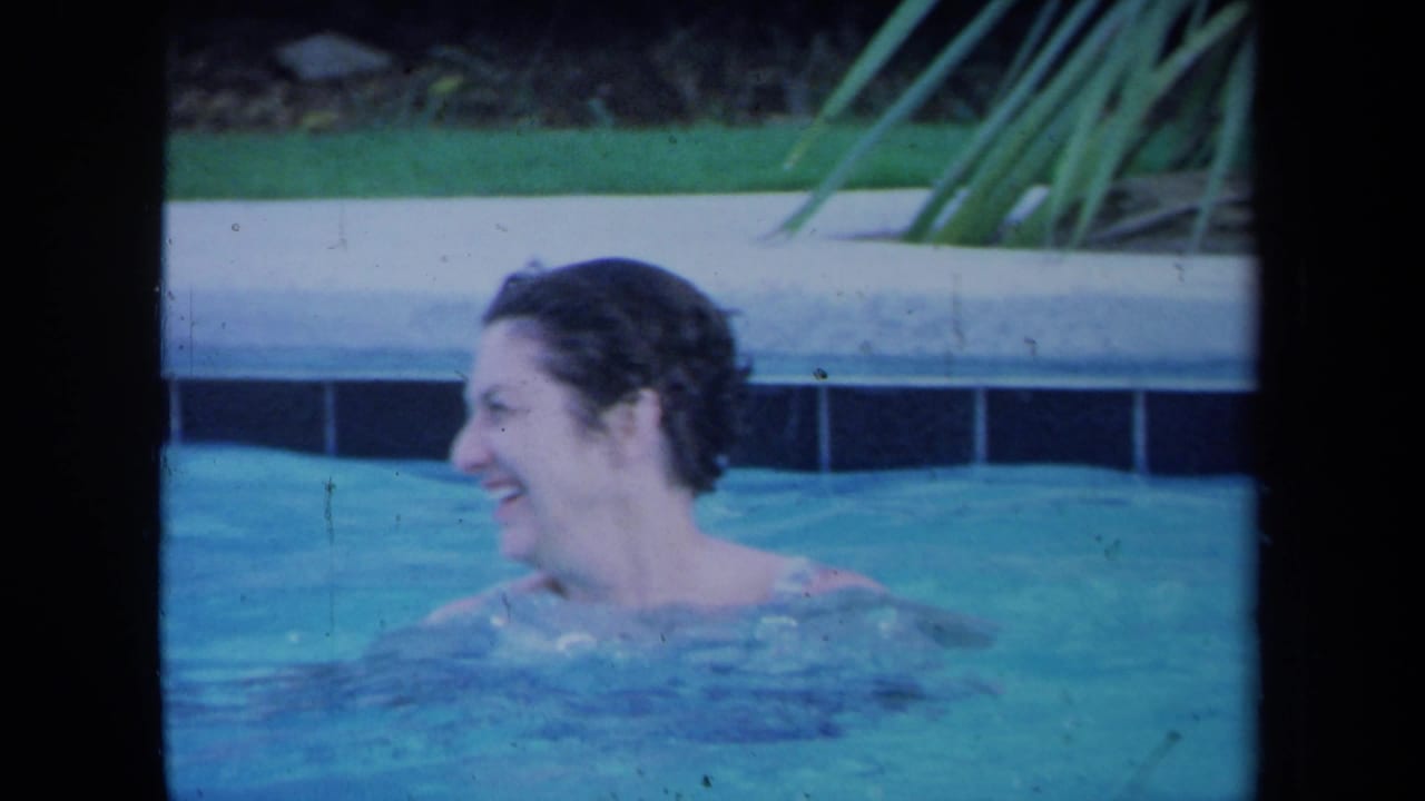 A woman smiles while turning in a backyard pool, with pool coping and greenery visible behind her in the 1971 California footage.