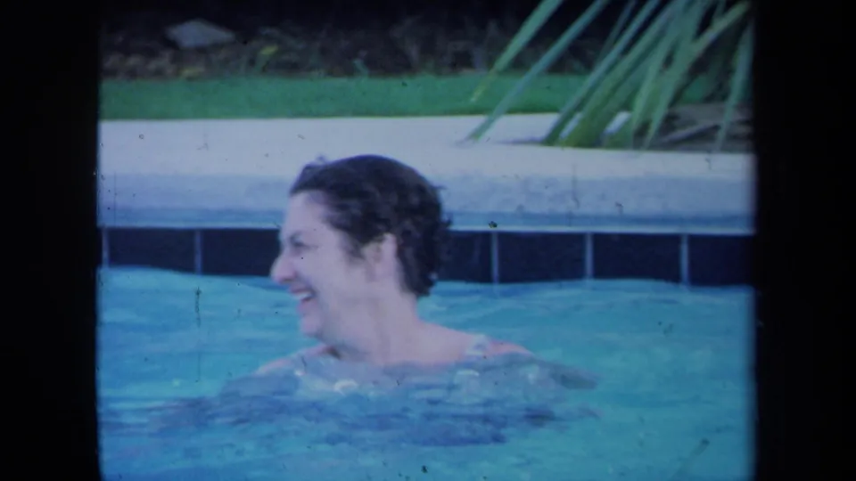 A woman smiles while turning in a backyard pool, with pool coping and greenery visible behind her in the 1971 California footage.