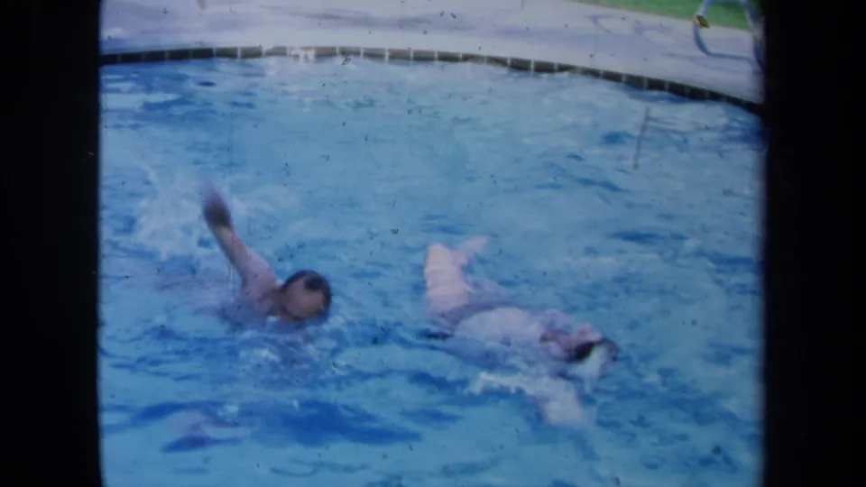 Two swimmers continue across an outdoor pool in 1971 California 8mm footage, with the pool edge visible at the top of frame.