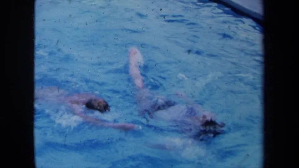 1971 California home-movie frame showing two swimmers in blue pool water with visible 8mm grain and film dust.