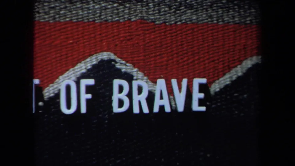 1971 California 8mm frame showing cropped white block letters “OF BRAVE” on a dark and red textured fabric surface.