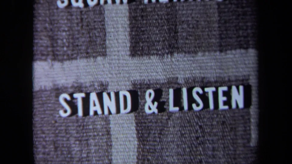 1971 California 8mm frame showing “STAND & LISTEN” in white block letters on a gray woven fabric background.