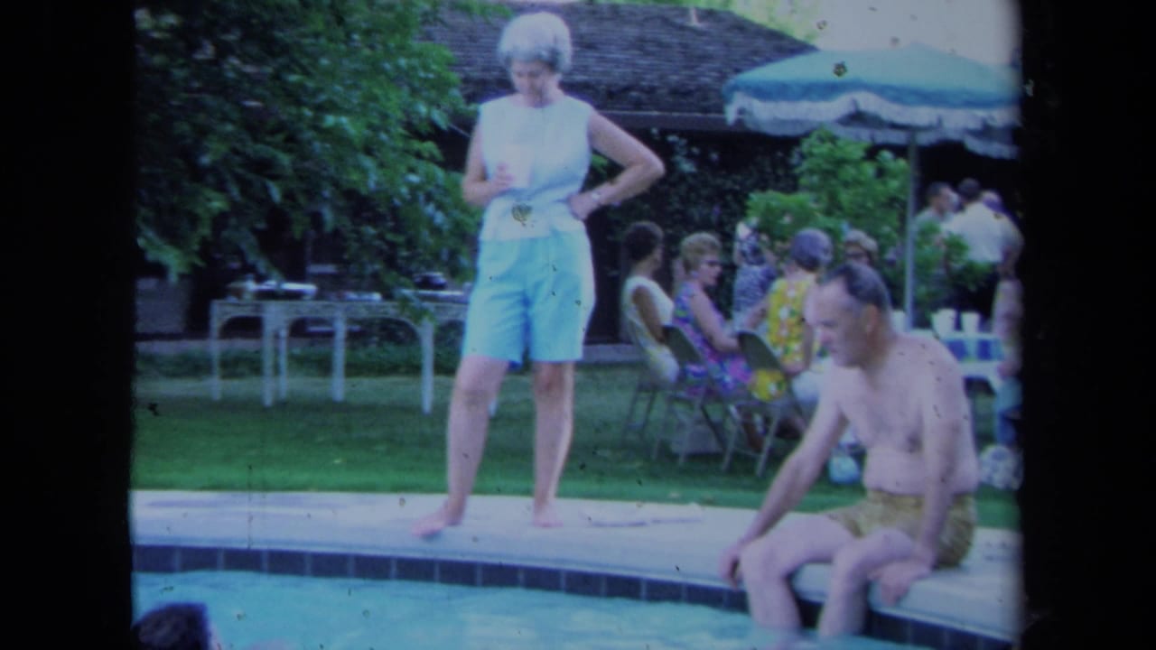 In 1971 California home-movie footage, adults socialize by a backyard pool while a woman stands at the edge and a man sits nearby.