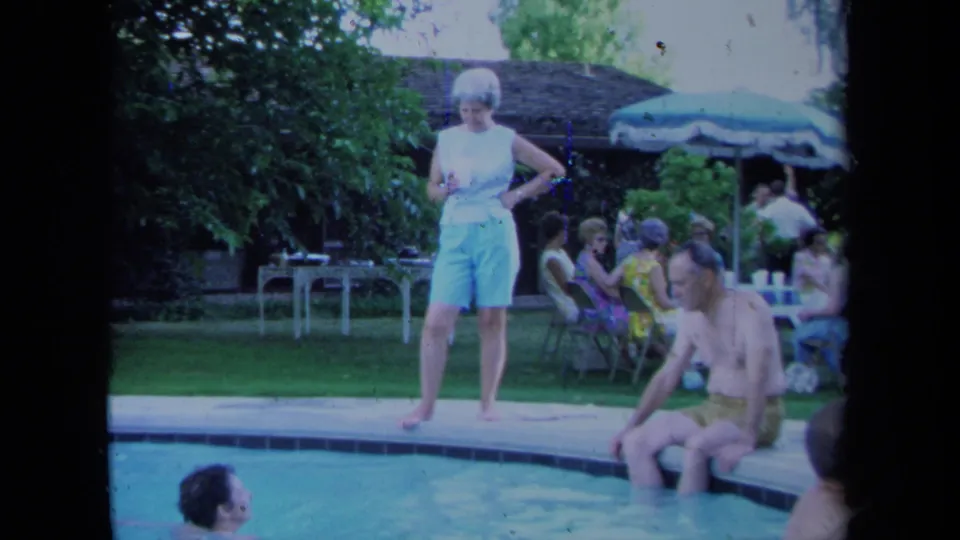 A woman in light clothing stands near the pool as a man sits at the edge and other adults talk on lawn chairs in the background.