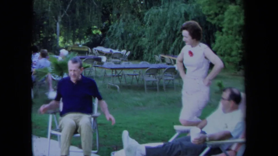 1971 California home-movie frame of adults gathered outdoors, with a seated man, a standing woman, and lawn furniture in the background.