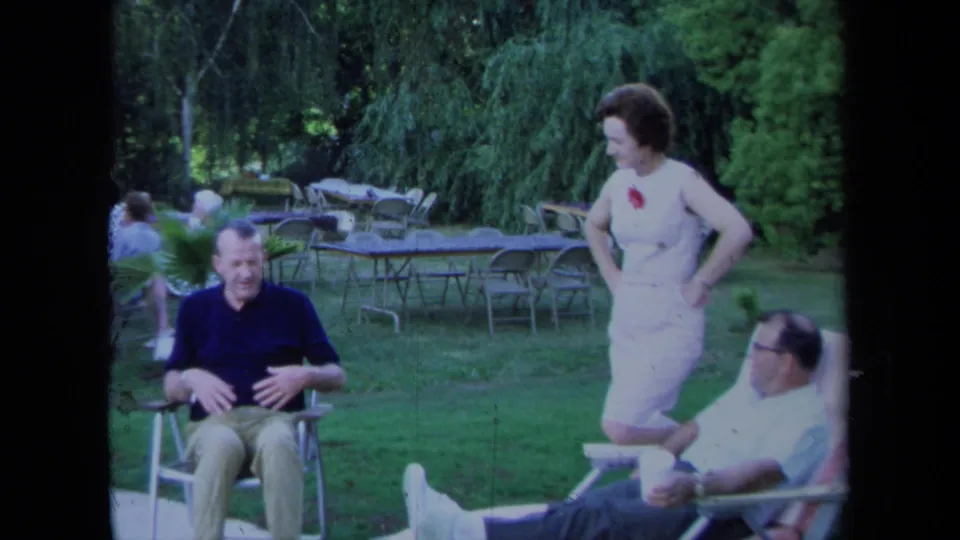 1971 California outdoor gathering with a seated man gesturing, a woman in a light dress standing, and another man reclining in a chair.