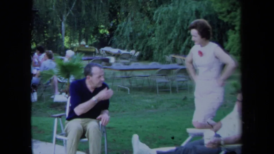 1971 California home-movie frame showing a seated man turning toward a standing woman during a casual lawn gathering.