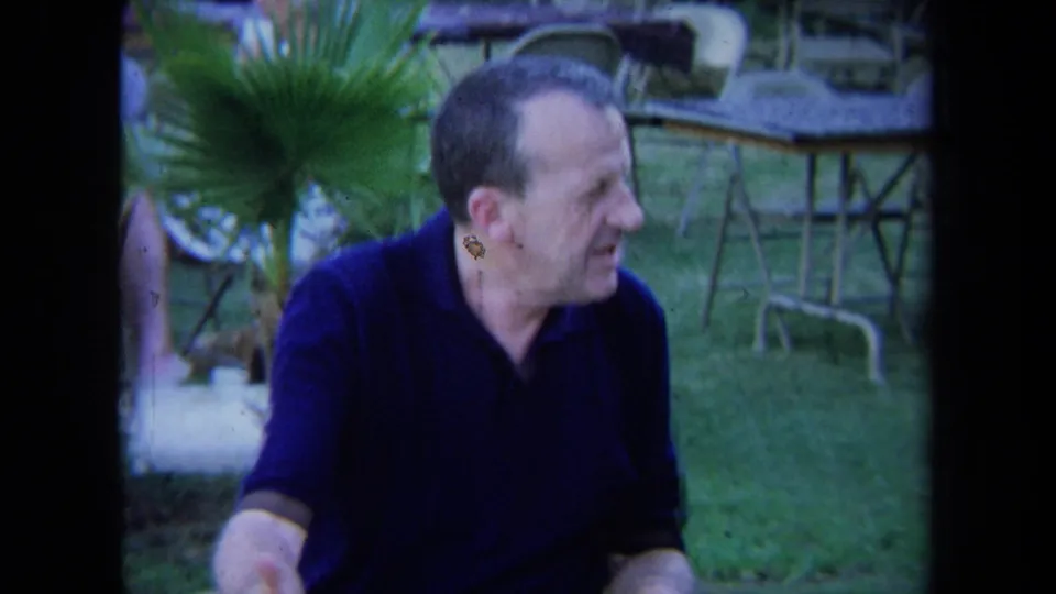 Close 1971 California home-movie view of a seated man in a dark polo, with grass, chairs, tables, and trees behind him.