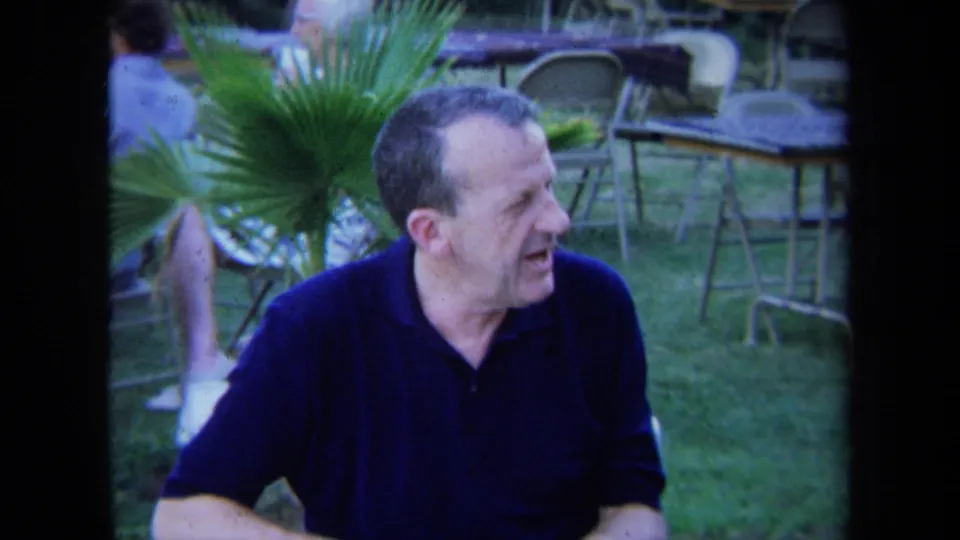 1971 California 8mm frame showing a middle-aged man in a dark polo shirt seated outside near lawn furniture and palm fronds.