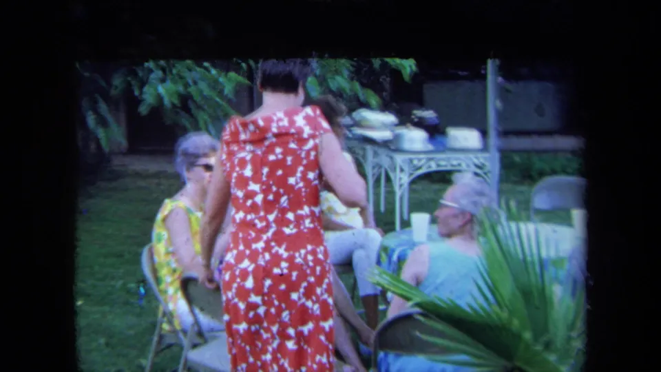 1971 California home-movie frame of women gathered on a lawn with a woman in a red and white patterned dress standing among chairs.
