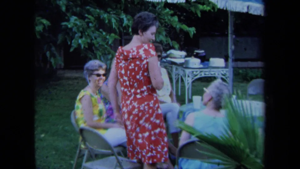 1971 California 8mm frame showing a woman in a red and white patterned dress among seated women on a lawn with refreshments behind them.
