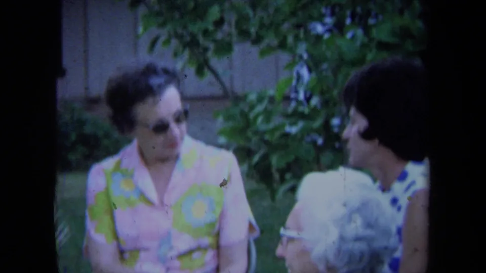 1971 California home-movie frame of older women conversing outdoors, with sunglasses, patterned clothing, and garden plants visible.