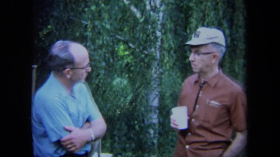 Two middle-aged men in 1971 California stand outside near dense greenery, one in a blue shirt with glasses and one in a brown shirt with a white cap holding a cup.