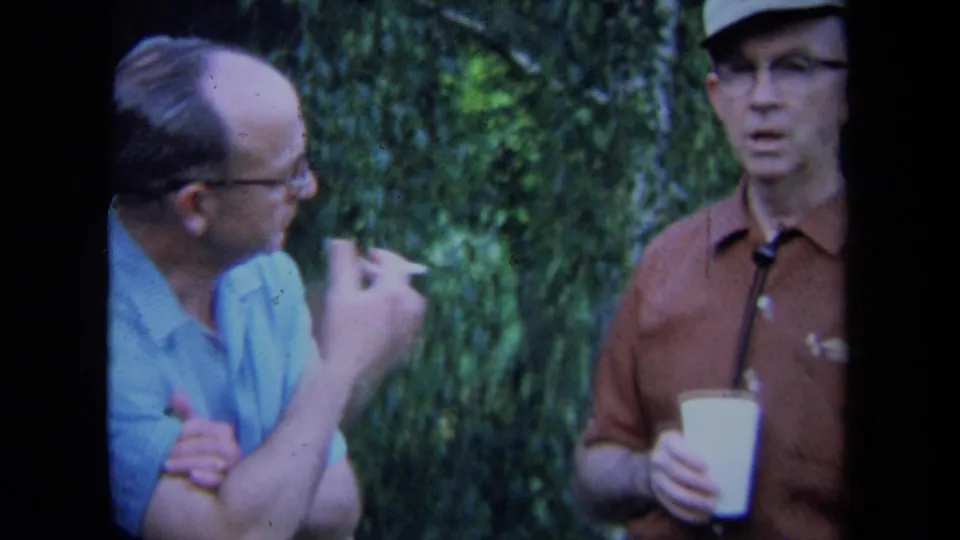 The two middle-aged men continue their outdoor conversation in California, with a cigarette visible and a cup held against a leafy background.