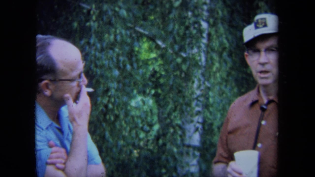 In a 1971 California home-movie frame, two adult men stand near dense greenery while one smokes and the other holds a cup.