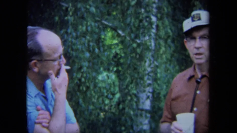 In a 1971 California home-movie frame, two adult men stand near dense greenery while one smokes and the other holds a cup.