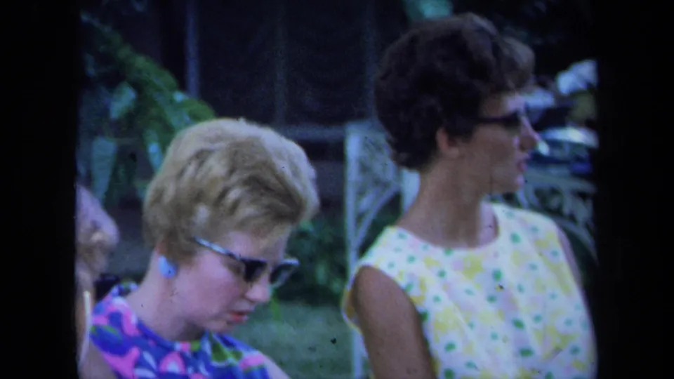 Two adult women appear in a garden, one with blonde hair and sunglasses and the other in a sleeveless floral dress.